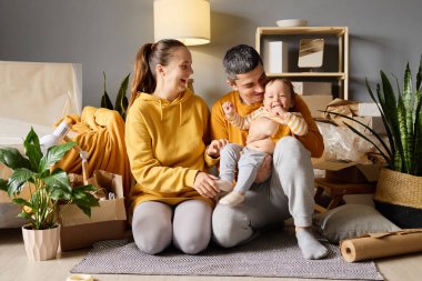 Mutlu genç ebeveynler yeni evlerinde neşeli bebekleriyle oynarken gülüyorlar. Etrafları kutularla çevrili, aileleriyle vakit geçiriyorlar..