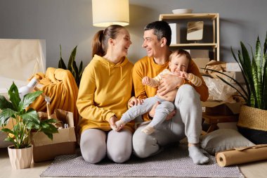 Mutlu genç ebeveynler yeni evlerinde neşeli bebekleriyle oynarken gülüyorlar. Etrafları kutularla çevrili, aileleriyle vakit geçiriyorlar..