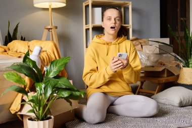 Şaşırmış genç bir kadın yeni evinde yerde oturuyor, etrafı kutularla çevrili, şok olmuş bir ifadeyle telefonuna bakıyor..