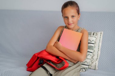 Nazik bir gülümsemeyle oturan genç bir kız pembe-turuncu bir not defteriyle kucağında kırmızı sırt çantasıyla sakince kendine güveni ve sıcaklığı yayıyor..