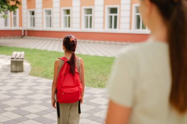 Kırmızı sırt çantalı genç bir kız okul bahçesinde dikilirken, ön planda bir kadının bulanık silueti görülüyor, bu da okulun bırakıldığını gösteriyor..