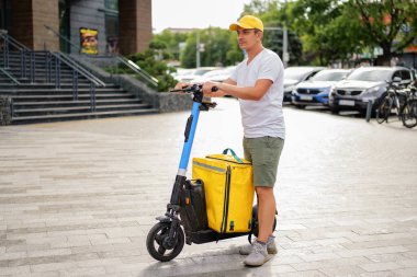 Sarı şapkalı bir teslimatçı, elektrikli scooterı ve büyük sarı bir teslimat çantasıyla şehir caddesinde duruyor..