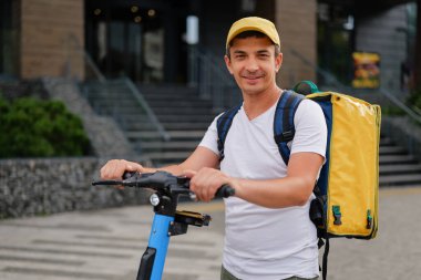 Sarı şapkalı, sarı çantalı gülümseyen bir teslimatçı, elektrikli scooterıyla bir şehir caddesinde duruyor..