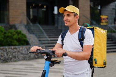 Sarı sırt çantalı ve beyzbol şapkalı gülümseyen bir teslimatçı şehir caddesinde e-scooterıyla dikiliyor..