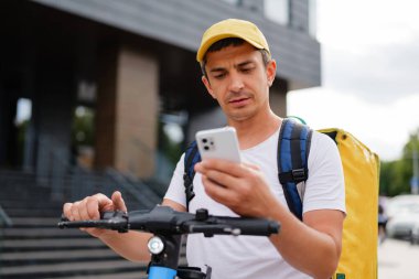 Sarı sırt çantalı ve şapkalı bir teslimatçı, şehir caddesinde e-scooterıyla dikiliyor, akıllı telefonuna odaklanmış bir ifadeyle bakıyor..