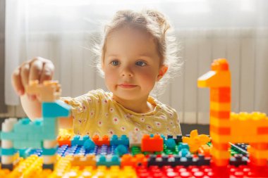 Renkli yapı taşlarıyla oynarken dağınık bir çörekle gülümseyen mutlu bir kız çocuğu, yeni bir şey yaratmaya odaklanmış..