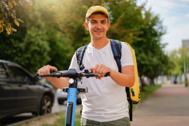 Sarı sırt çantalı gülen kurye güneşli bir günde elektrikli scooter kullanıyor. Hızlı ve modern teslimat servisi.
