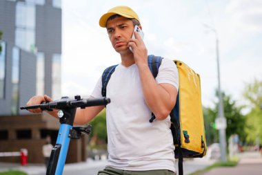Sırt çantalı bir teslimatçı telefonuyla konuşuyor, ciddi bir ifadeyle, bir müşteriyi ya da hareket memurunu dinliyor..