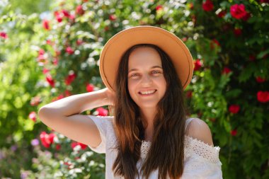 Hasır şapkalı ve beyaz elbiseli gülümseyen genç bir kadın güneşli bir bahçede kırmızı çiçekli bir portre için poz veriyor..