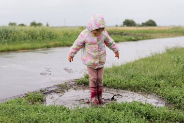 Yağmurluk ve çizme giymiş genç bir kız yeşil bir tarlada çamurlu bir su birikintisinde duruyor, neşeyle suya bakıyor..
