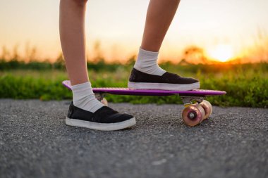 Güzel bir gün batımında, asfalt bir yolda mor bir kaykayın üzerinde duran bir insanın bacak ve ayaklarının yakın çekimi..