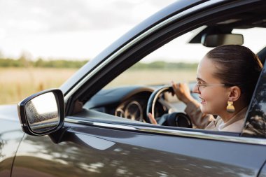 Mutlu bir iş kadını arabasını sürerken gülümser. Manzara yandan, ön planda dikiz aynası var..