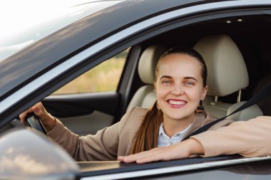 Bej takım elbiseli arkadaş canlısı bir iş kadını arabasının camından dışarı bakıyor ve kameraya gülümsüyor..