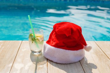 Neşeli bir Noel Baba şapkası, berrak mavi gökyüzünün altında, köpüklü bir havuzun kenarında, soğuk bir içecek bardağının yanında duruyor..