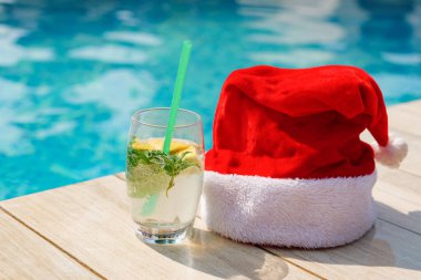 Limon ve nane aromalı renkli bir kokteyl parlak gün ışığı altında parlak bir havuz altında ahşap bir Noel Baba şapkasının yanında oturur..