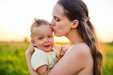 Bir kadın, gün batımında onu canlı yeşil bir tarlada tutarken nazikçe gülümseyen bebeğini öper. Yumuşak, altın ışıkla çevrili..
