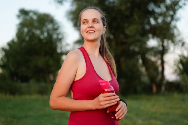 Kırmızı aktif giysili neşeli ve formda bir kadın, güneşli bir parkta spor yaptıktan sonra elinde su şişesiyle gülümsüyor..