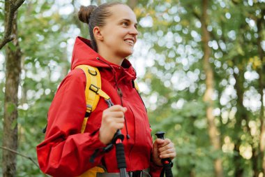 Sarı bir sırt çantası ve yürüyüş direkleri olan neşeli bir kadın canlı yeşil bir ormanda yürürken gülümsüyor..