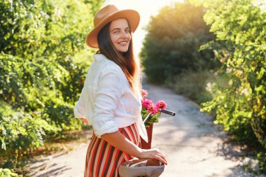Gülen kadın şapkalı bisiklet ve çiçeklerle dolu sepetin içinde yeşil bir yolda yürüyor..