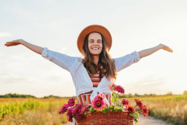 Kollarını uzatmış neşeli bir genç kadın kırsalda güneşli bir günde elinde bir sepet çiçekle klasik bir bisiklete biniyor. Şapka, beyaz gömlek ve çizgili bir elbise giyiyor..