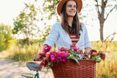 Çizgili elbiseli ve şapkalı neşeli genç bir kadın kırsal kesimde güneşli bir günde, canlı çiçeklerle dolu bir sepeti olan antika bisikletinin yanında duruyor..