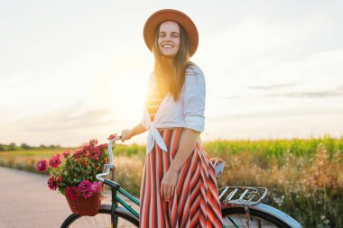 Çizgili elbiseli ve şapkalı gülümseyen bir kadın elinde bir sepet çiçekle klasik bisikletiyle kırsalda sıcak ve güneşli bir günün tadını çıkarıyor..