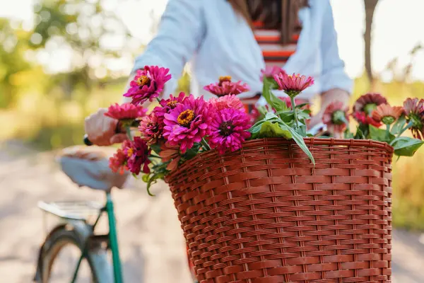 Bir sepet dolusu canlı pembe ve mor çiçekli antika bir bisikletin yakın plan fotoğrafı. Arka planda odak dışı bir kişi görülüyor.