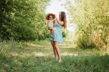 Gülümseyen bir anne, güneşli bir günde bir parkta şapka takan mutlu küçük oğlunu kucağına alır..