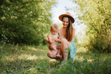 Güneşli bir günde çimenli bir parkta, kucağında küçük oğluyla çömelmiş, oynaşan, yaz elbisesi içinde mutlu bir anne..