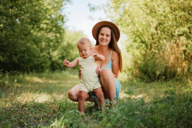 Güneşli bir günde çimenli bir parkta ilk adımlarını atan küçük oğlunun yanına çömelmiş güneş elbisesi giymiş genç bir anne..