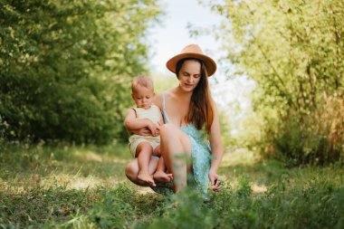 Yazlık bir elbise içinde gülümseyen genç bir anne, güneşli bir günde çimenli bir parkta kucağında oturan küçük oğluyla çömeliyor ve oynuyor..