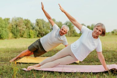 Parkta yaşlı bir çift yoga minderleri üzerinde yan tahtayı geriyor..