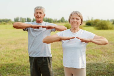 Aktif kıdemli çift sahada dikiliyor ve sağlıklı yaşam tarzlarını temsil eden yatay bir kol esnemesi yapıyor..