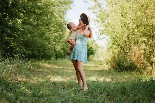 Güneşli bir günde parkta gülümseyen ve birbirine bakan, küçük oğlunu kollarında tutan mutlu bir anne..