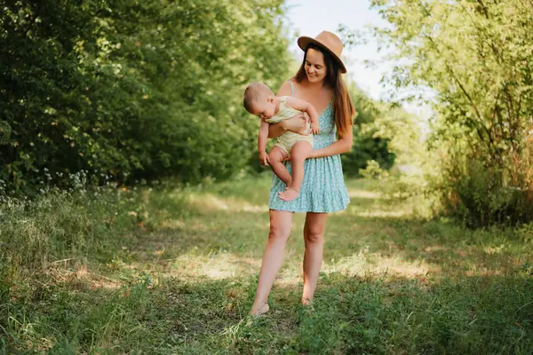 Yazlık elbiseli ve şapkalı genç bir anne, güneşli bir günde çimenli bir tarlada dikilirken, bir gülümsemeyle küçük oğlunu kucağına alır..