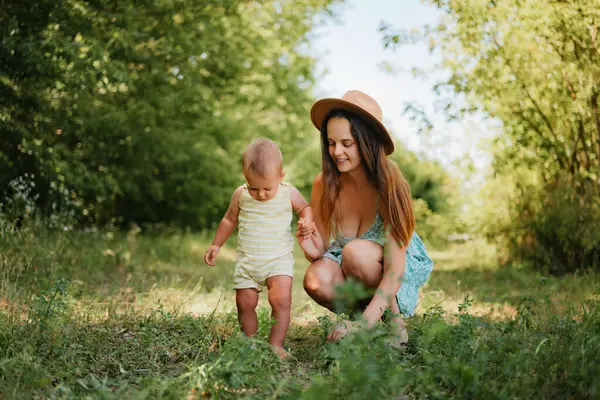 Güneşli bir günde çimenli bir parkta ilk adımlarını atarken küçük oğlunun elini tutan yaz elbisesi içinde gülümseyen genç bir anne..