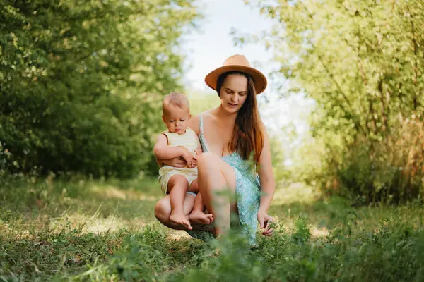 Yazlık bir elbise içinde gülümseyen genç bir anne, güneşli bir günde çimenli bir parkta kucağında oturan küçük oğluyla çömeliyor ve oynuyor..
