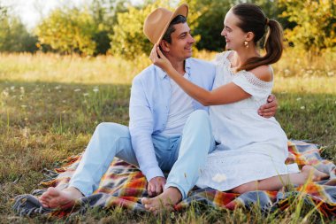 Mutlu bir çift, tarlada bir battaniyenin üzerinde romantik bir pikniğin tadını çıkarıyor, gülümsüyor ve birbirlerine bakıyorlar..
