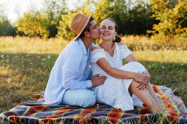 Romantik bir çift pikniğin tadını çıkarırken, erkek, kadın gözleri kapalı gülümserken yanağından öpüyor.