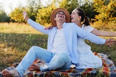 Gülen bir çift, tarlada battaniyede pikniğin tadını çıkarıyor, kolları açık bir şekilde..