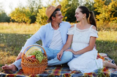 Piknik battaniyesinde neşeli bir çift, birbirlerine bakıyor ve aralarında bir sepet taze meyveyle gülümsüyorlar..