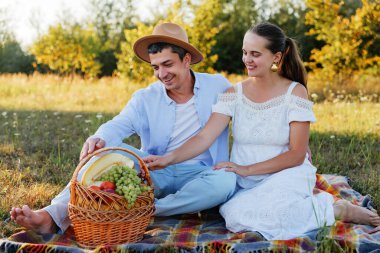 Piknik battaniyesi üzerinde gülümseyen bir çift, aralarında meyve sepetine uzanıyorlar..