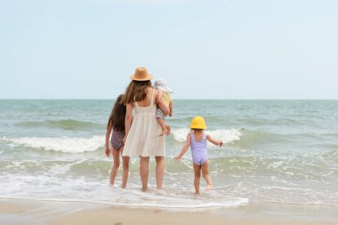 Bir anne, güneşli bir sahil gününde hafif dalgalar ayaklarına vururken çocuklarının yanında durur..