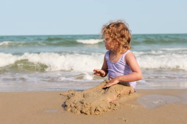 Gün ışığında parlayan parlak buklelerle bir çocuk, sıcak bir sahilde oynarken dalgaların kenarında kumdan kale inşa etmekten zevk alır..
