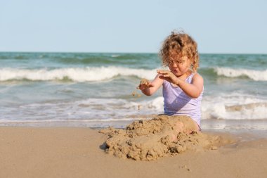 Genç bir kız kumlu sahilde oturuyor, kumdan kale inşa etmeye odaklanmış dalgalar açık mavi gökyüzünün altında yavaşça süzülürken.