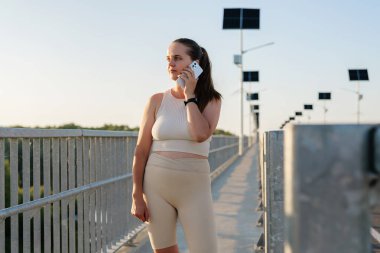 Aktif giyimli bir kadın, parlak bir yolda duruyor. Altın saat boyunca güneş ışığıyla çevrili bir telefon görüşmesi yapıyor..