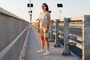 Genç bir kadın, gün batımında sıcak güneşin ve dingin atmosferin tadını çıkarırken, idman kıyafetlerini giymiş, güven içinde köprüde duruyor..