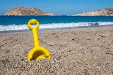 Plajda oynamak için kullanılan sarı plastik kürek kalp şeklinde sapıyla turkuaz deniz dalgalarının önünde kumların üzerinde duruyor.