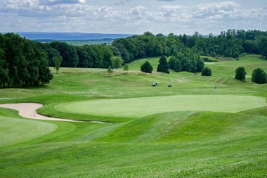 Golf sahasının pürüzsüz çimenli bölgesi, ağaçlar ve bulutlu gökyüzü.