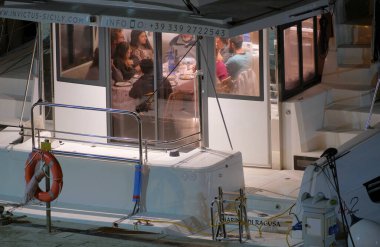 İtalya, Sicilya, Akdeniz, Marina di Ragusa (Ragusa Eyaleti); 17 Aralık 2022, insanlar geceleri limandaki bir katamaran yelkenli teknesinde yemek yiyorlar -